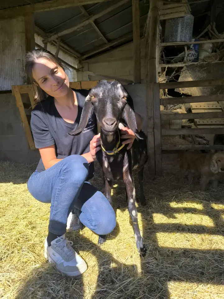 Marine Boullier souriante, accroupie dans la paille, entourant de son bras une chèvre tachetée aux longues oreilles